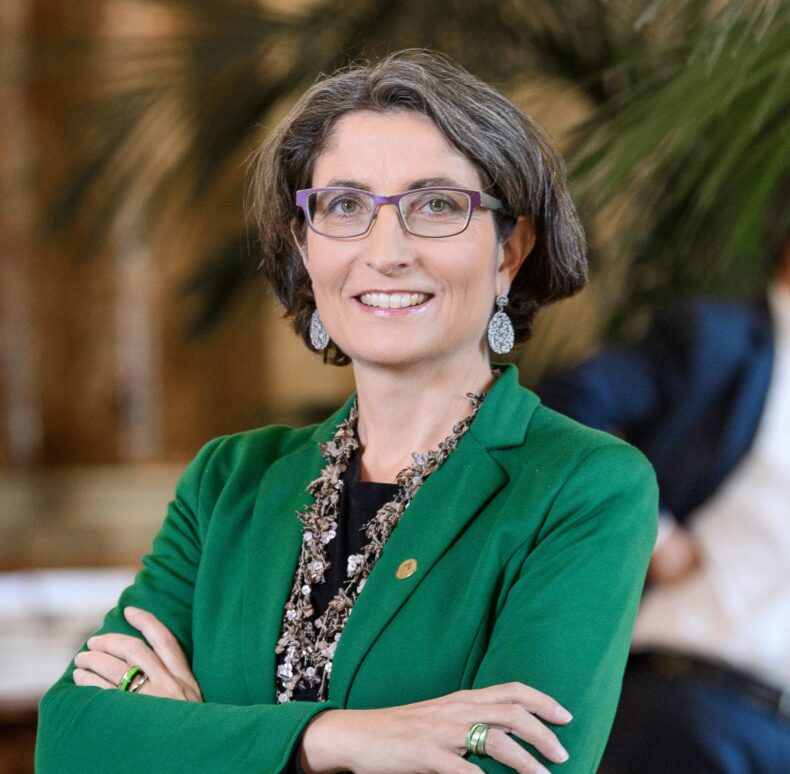 Manuela Weichelt in einem noblen grünen Anzug, einer violetten Brille, gepflegter Frisur, lächelnd, grosse ovale Ohrenhänger bestehend as vielen kleinen Perlen drauf, spannende Kette aus Steinen und weiteren Nautrgegenständen.